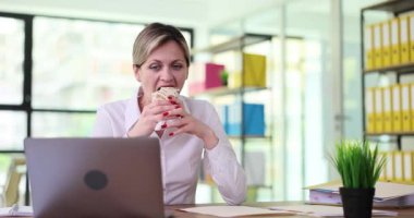 İş kadını yemek molasında ofiste dürüm yiyor ve televizyon seyrediyor. Doğal gün ışığı çalışma alanını doldurur ve rahat bir atmosfer yaratır.