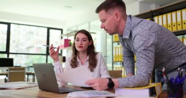 Kadın elinde kalem tutarken adam ekranı gösteren basılı belgeyi açıklıyor. Gün ışığı ofise giriyor üretken bir atmosfer yaratıyor