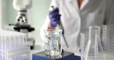 Laboratuvar önlüklü bilim adamı deney kabında dönen sıvının yanında pipet kullanıyor. Girdap, kimyasal deney sırasında hızla döner ve dinamik hareket yaratır.