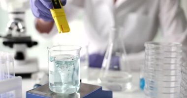 Laboratuvar teknisyeni deney sırasında sarı pH testçisini girdap sıvısına batırıyor. İşçi el pozisyonunu ayarlayan ölçümü okuyor