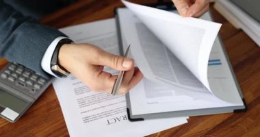 Takım elbiseli bir işadamı kalem kullanarak sözleşme sayfalarını çeviriyor. Parlak ışık ofise girer çalışma sırasında ahşap masa yüzeyinde kontrastlık yaratır