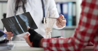 Kadın doktor, klinikte bilek röntgeninde ve avuç içi kemiklerinde yaralanma olduğunu gösteriyor. Elindeki bandajlı hasta randevudaki doktoru dinliyor.