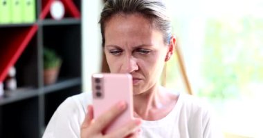 Beyaz tişörtlü kadın gergin bir ifadeyle pembe akıllı telefonu inceliyor. Yaralı kaşlar ve odaklanmış bakışlar görme sorunları olduğunu gösteriyor.