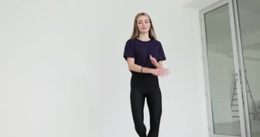 Siyah tişört giyen ve tayt giyen bir kadın beyaz duvarlı, parlak minimalist bir odada dans ediyor. Kollar hafifçe kaldırılmış ve bükülmüş aktif hareketleri gösterir