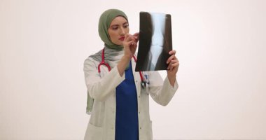 Beyaz önlüklü ve yeşil tesettürlü doktor röntgenleri dikkatle inceler. Kalemi olan ciddi bir kadın radyografa ilgi alanlarını işaret ediyor.