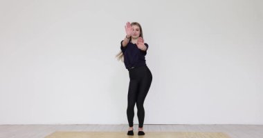 Genç bir kadın dans pozu veriyor. Kolları açık, avuçları dışarı bakıyor. Minimalist iç mekanda odaklanmış ve kendinden emin ifade