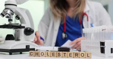 Tahta bloklar mikroskobun yanında kolesterol ve klinik masasında laboratuvar tüpleri olduğunu gösteriyor. Doktor kan örneklerini inceler ve kalp riski hakkında notlar yazar.