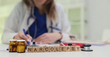 Tahta harfler kahverengi ilaç kavanozlarının yanında duran narkolepsi kelimelerini heceliyor. Uyku uzmanı masada oturur ve uyku bozukluğu tedavisi planı için notlar alır.