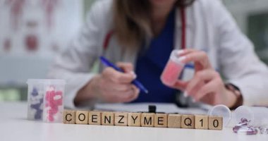 Tahta harfler, vitaminlerin yanındaki beyaz yüzeyde duran Koenzim Q10 'u heceliyor. Laboratuvar önlüklü kadın hasta için reçeteli ilave bilgi ekliyor