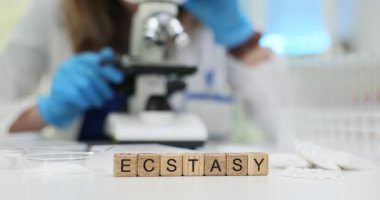 Tahta küplerde mikroskop ve laboratuvar aletlerinin yanında duran Ecstasy kelimesi hecelenir. Toz örnekleri ve mühürlü paketler steril ortamdaki uyuşturucu analizini ortaya koyuyor