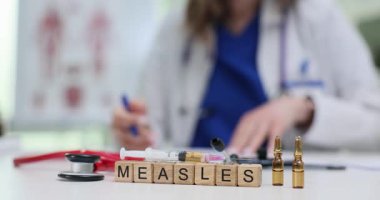 Doktor tahta bloklarda kızamık kelimesinin yanında kalem ve belge tutuyor. Tıbbi araçlar, klinikteki bulaşıcı hastalıklara ve aşılara odaklandığını gösteriyor