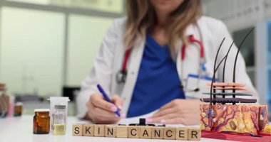 Doktor, masasındaki tahta kiremitlere cilt kanseriyle ilgili belgeler yazıyor. İnsan derisi ve saç foliküllerinin anatomik modeli dermatolojiye odaklandığını gösteriyor.