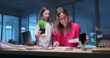 Genç kız, masa başına saksıları yerleştirip evrak işlerine odaklanmış bir kadını rahatsız ediyor. Yorgun anne, kızı koşarken gözlerini deviriyor.