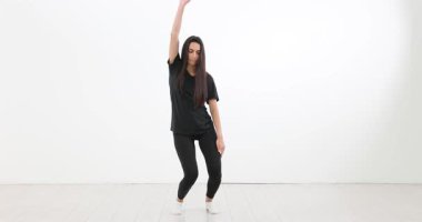 Siyah eşofmanlı zayıf, uzun saçlı kadın stüdyoda dans ediyor. Kaygısız esmer kadın, beyaz arka planda ağır çekimde vücut hareketleri yapıyor.