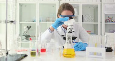 Mutlu bayan teknisyen mikroskoba bakar ve laboratuvarda el sallar. Kadınlar laboratuvar incelemeleri sırasında yavaş çekimde selamlama hareketi gösteriyor.