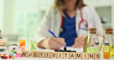 Doktor ahşap blokların yanında dokümanlar imzalar. Masasında haplar ve şişelerle birlikte Multivitaminler bulunur. Sağlık desteğine ve tıbbi danışmaya odaklanın