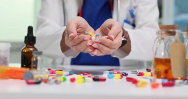 Doktor, beyaz yüzeye yayılmış ilaç şişeleriyle büyük miktarda renkli haplar gösteriyor. Sahne güçlü ilaç temaları taşır