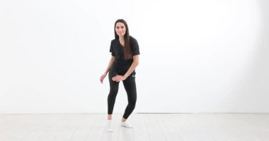 Siyah kıyafetli kadın dinamik danslar yapıyor ve beyaz duvarda egzersiz yapıyor. Stüdyo çekimi hareket ve fiziksel ifade üzerine yoğunlaşır.