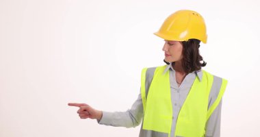 Sarı kasketli ve yüksek görüşlü yelekli bir kadın işaret parmaklarıyla gülümsüyor. Yön kavramı ve inşaattaki önemli alan