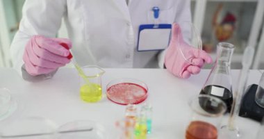 Bilim adamı pipeti kavanozun üstünde tutuyor. Petri kabına hassasiyetle sarı sıvı ekliyor. Parmaklar, kimyasal teması kontrol eden bir kap.