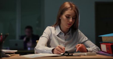 Ofisteki genç bayan hesap makinesi kullanarak not defterine hesaplamalar yapıyor. İş kadını gece geç saatlere kadar ofiste kağıt üzerinde bütçe işleriyle uğraşır.