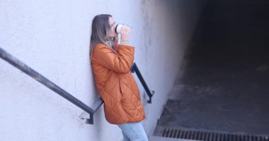 Turuncu ceketli kadın tünelin yanında elinde kahve fincanı tutan tırabzanlara yaslanıyor. Kadın uzaklara bakar ve karanlık yeraltı geçidini dikkatle izler.