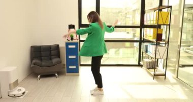 Neşeli iş kadını modern ofiste neşeli bir şekilde dans ediyor. Parlak yeşil ceketli kadın ofis çalışanı bulaşıcı enerjiyle kollarını hareket ettiriyor.