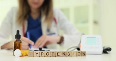 Laboratuvar önlüklü kadın, tahta bloklarda hipotansiyonlu notlar alıyor. Tansiyon monitörü düşük tansiyon teşhisi ve yönetimi öneriyor.