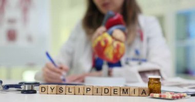 Küpten yapılan Dyslipidemia kelimesi kapsüllerin ve anatomik kalp modelinin yanında duruyor. Kadın hasta kolesterol dengesizliğini belgeleyen notlar alıyor.