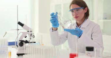 Genç bayan laboratuvar asistanı, damıtılmış suyla dolu şişeyi dikkatlice eğiyor ve berraklığı inceliyor. Kadın sıvı hareketini izler. Yavaş çekim