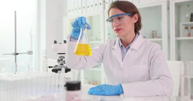 Kadın kimyager sarı çözeltiye sarılır ve değişiklikleri gözlemler. Dişi matarayı göz hizasına yükseltir ve sıvının renk ve hareketini kontrol eder. Yavaş çekim