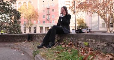 Esmer kadın telefonda el hareketleriyle konuşuyor. Parktaki taş çıkıntıda oturuyor. Sonbahar yaprakları duygusal iletişim havası yaratıyor.