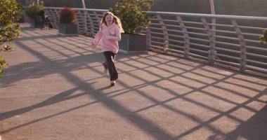 Pembe kazaklı genç kız gün batımında yaya köprüsünden geçiyor. Korkulukların gölgeleri kaldırıma doğru uzanıyor. Enerjik bir ritm oluşturuyor. Yavaş çekim.