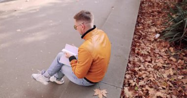 Turuncu ceketli adam sonbahar parkının korkuluklarında oturur ve defter okur. Düşen yapraklar mevsimsel bir ruh hali yaratır.