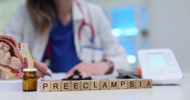 Tahta küplerden oluşan preeklampsi kelimesi rahim modelinin yanında ve doktor masasında kan basıncı monitörünün yanında duruyor. Haber hamilelik komplikasyonunu yansıtıyor.