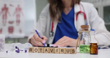 Tahta bloklar Rotavirüs kelimesinin renkli kapsüllerin ve laboratuvar şişelerinin yanında durduğunu söyler. Doktor rotavirüs enfeksiyonu ve aşı üzerine notlar yazıyor.