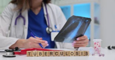 Tahta bloklar tüberküloz kelimesinin pano ve kalemin yanında durması anlamına geliyor. Doktor akciğer röntgeni görüntüsünü inceleyip tüberküloz çalışması için notlar yazıyor.