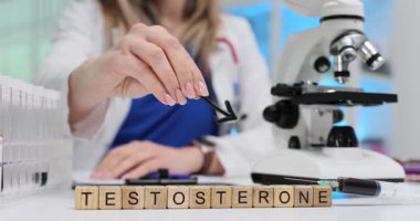 Tahta bloklar laboratuvar mikroskobu ile yerleştirilen testosteron kelimelerini heceler. Doktor okları mikroskoba doğrultup örnek analizini ağır çekimde sembolize ediyor.