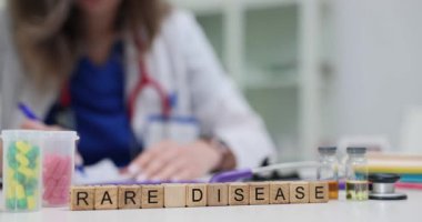 Tıp önlüklü kadın hasta geçmişini nadir görülen hastalıklar ve masasında haplar olan küplerle yazıyor. Bayan nadir görülen hastalıkları teşhis etmeye ve tedavi etmeye odaklanıyor.