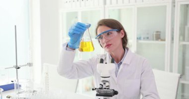 Kadın laboratuvar asistanı test tüpünde sarı sıvıyı dikkatle gözlemliyor. Laboratuvar çalışanı reaksiyon sürecini belirlemek için matarayı inceliyor. Yavaş çekim