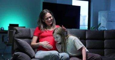 Kız hamile kadının karnına yaslanıp bebek hareketlerini dinliyor. Kanepede gülümseyen bir kadın büyük kızıyla nazik bir anın tadını çıkarıyor.