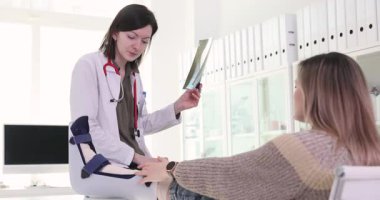 Bacağı alçılı bir kadın, doktorun dinlenme topuğunu masada ziyaret ediyor. Radyoloji uzmanı, görüntüleme sonucu ve tedavi adımlarını tavsiye eden uzuvları inceliyor