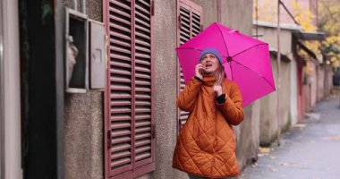 Pembe şemsiye altındaki gergin kadın, sonbahar caddesinde ayakta duran akıllı telefondan konuşuyor. Yaşlı bir kadın, yağmurlu havadan şikâyet ediyor.