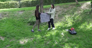 Kadınlar piknik için rahat bir yer hazırlamak için yeşil çimenlere geniş battaniye sererler. Kız arkadaşlar battaniyeyi uzatıyorlar oturulacak yeri rahatlatmak için.