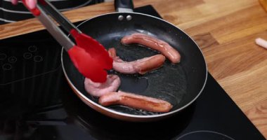 Elinde maşa olan kadın mutfaktaki tavada sosisleri çeviriyor. Kadın, öğle yemeği için evde ağır çekimde tümevarımlı fırında et kızartıyor.