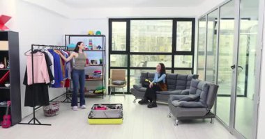 Kadın, stüdyo dairesinde kız arkadaşıyla seyahat etmek için kapüşonluyu seçiyor. Genç bayan eşofman üstünü bavula yerleştirirken, yol arkadaşı kanepeye oturur.