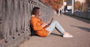 Serbest çalışan bir kadın, sonbahar günü dizüstü bilgisayarla şehir yolunda oturup çitlere yaslanıyor. Sunny Park 'taki uzaktan kumandalı bayan çalışan portatif cihazda çalışıyor.