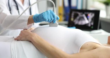 Doktor, hasta yatağında rahat bir şekilde yatan hamile bir kadın için ultrason muayenesi yapıyor. Doktor randevuda doğmamış bebeğin gelişimini kontrol ediyor.