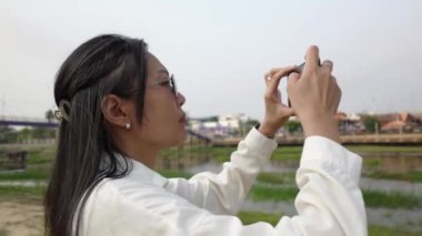 Asyalı bir kadın cep telefonuyla nehir kenarında fotoğraf çekiyor.