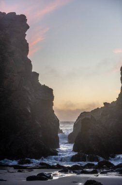 Gökyüzünde turuncu bulutlarla Pfeiffer Sahili 'ndeki kayalıkların arkasında batan güneş..